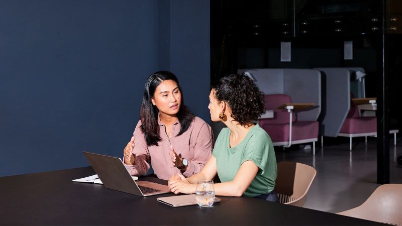 Two women sitting at desk with computer having discussion