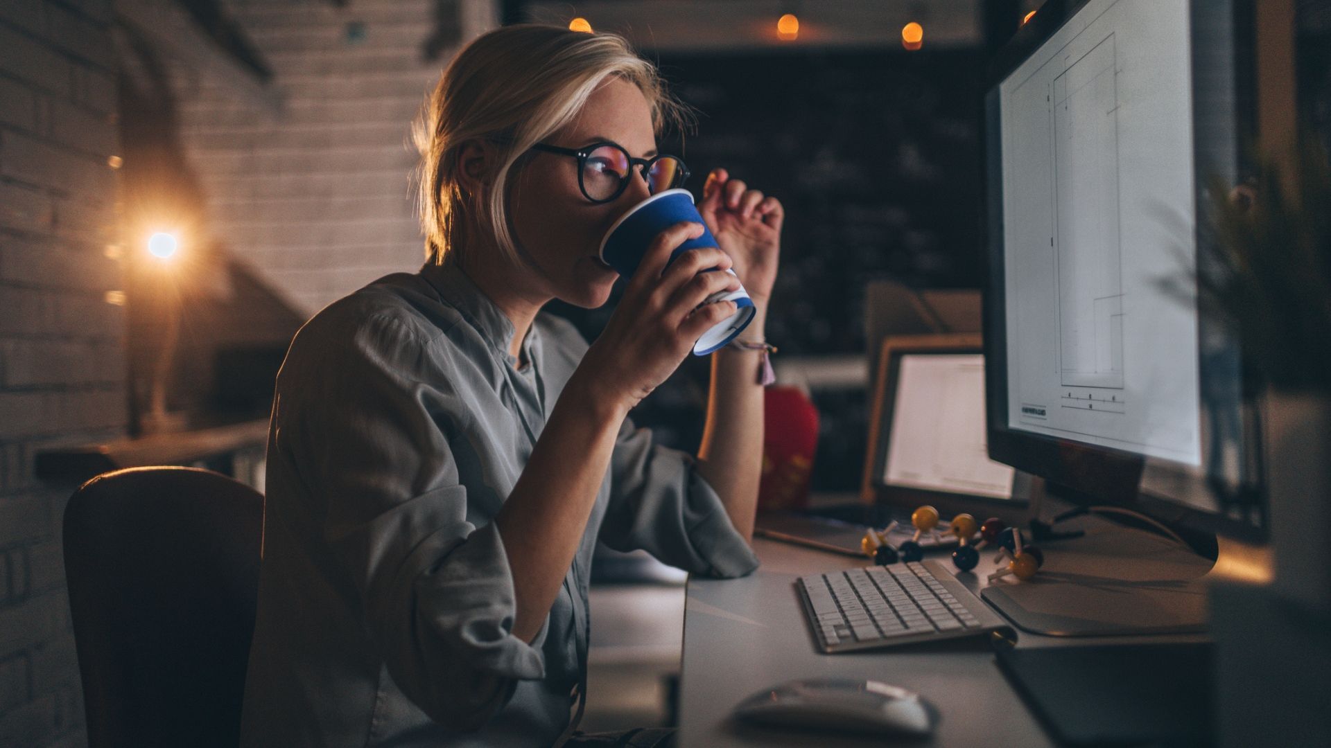 Young woman sitting at her desktop computer having some coffee to help her through a late work night. 