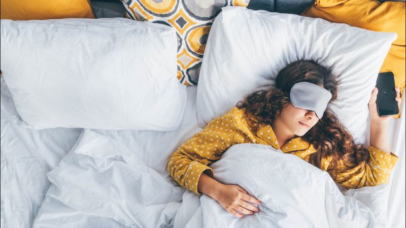 Woman lying in bed with sleeping mask and phone on her hand