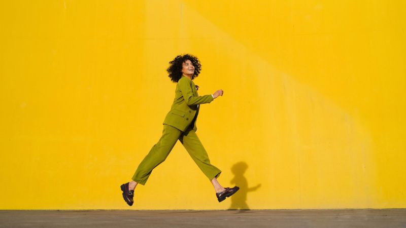 Energetic woman in business attire leaping against bright yellow urban backdrop