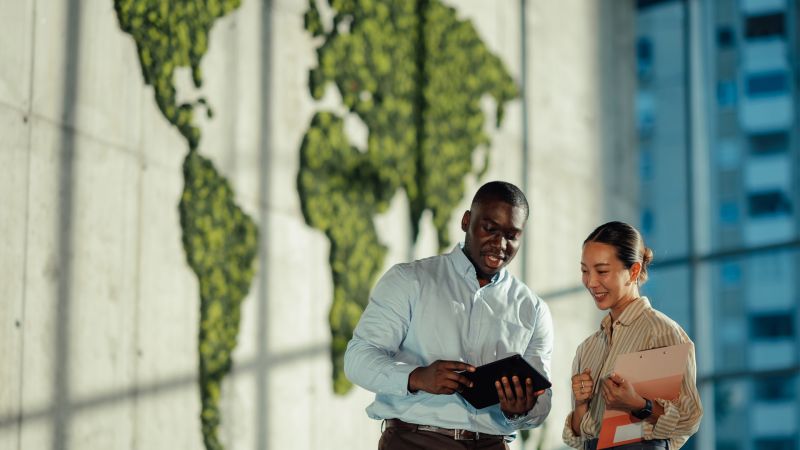 Two managers are discussing sustainable business strategies using digital tablet and clipboard, with a green world map in the background symbolizing global environmental awareness
