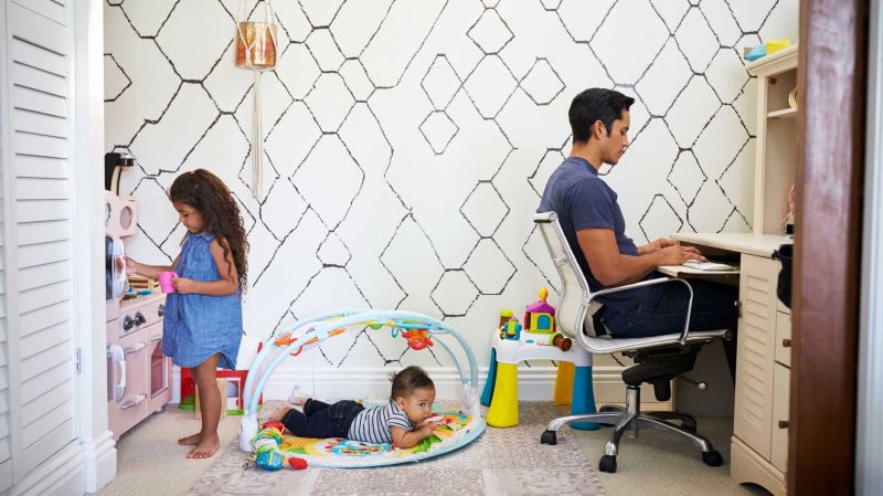 Dad sits working at a desk at home while his baby son and young daughter play in the room behind him.