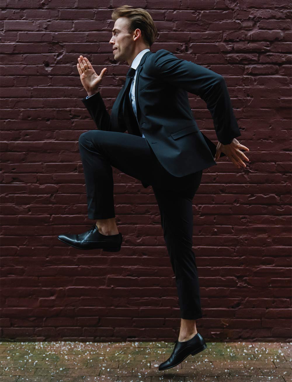 Connor Emery is a man in a nice suit with styled brown hair. He is suspended in the air in a running pose in front of a brick wall.