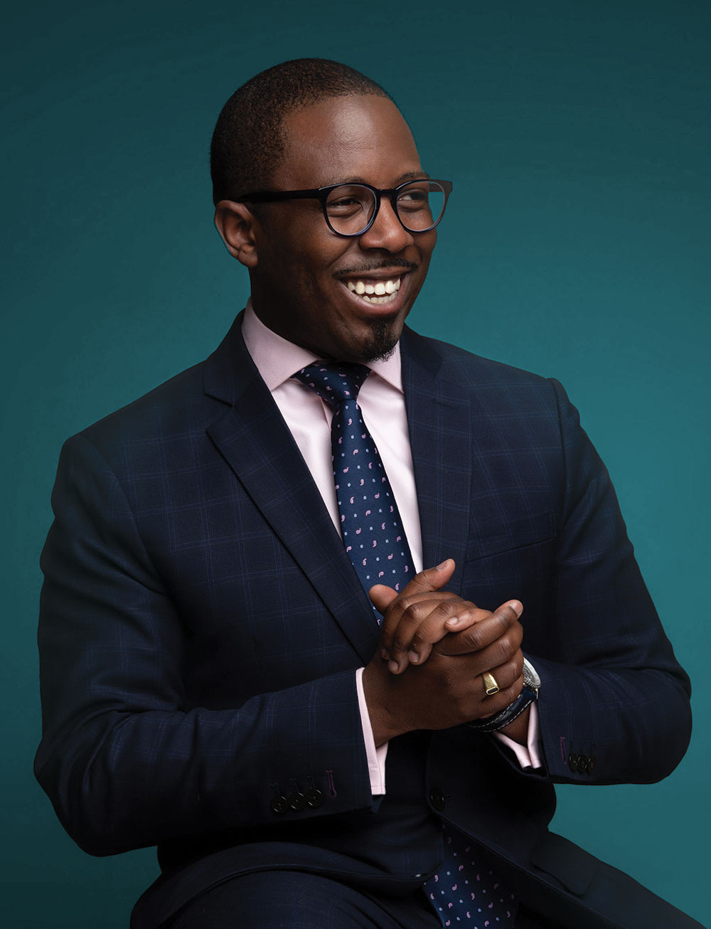 Aziz Garuba is a smiling dark skinned man sitting in front of a turquoise background.