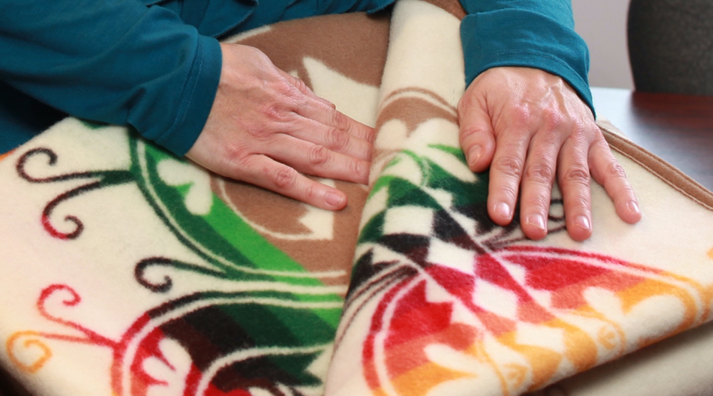 A person’s hands over an Indigenous blanket that is white, with green, red and yellow accent colours